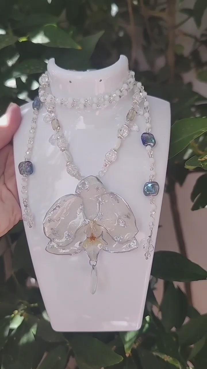 Collar largo hecho a mano con orquídea real blanca resinada, detalles plateados y corazón de piedra luna
